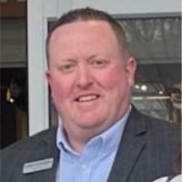 a man wearing a black and grey suit with a blue undershirt unbuttoned. He has a grey name tag on that is not readable. 