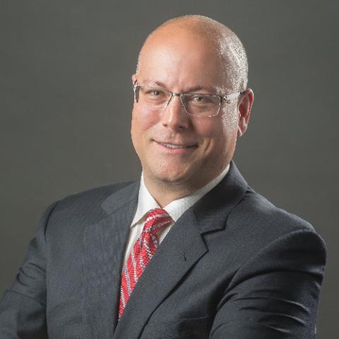 a headshot of a bald man with glasses in a black suit with a red tie and a white shirt against a dark grey background