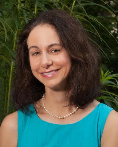 A woman with dark hair, blue dress, and pearls against a greenery background