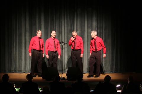 four men singing against a black backdrop with red shirts and black pants