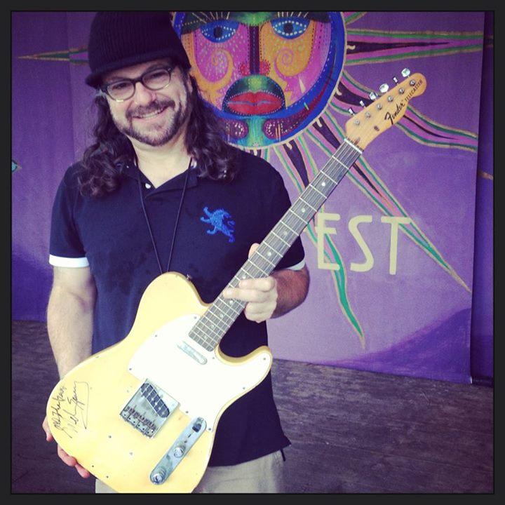 Joey Leone holding a Yellow Guitar against a purple background. The picture has a lens vignetting 