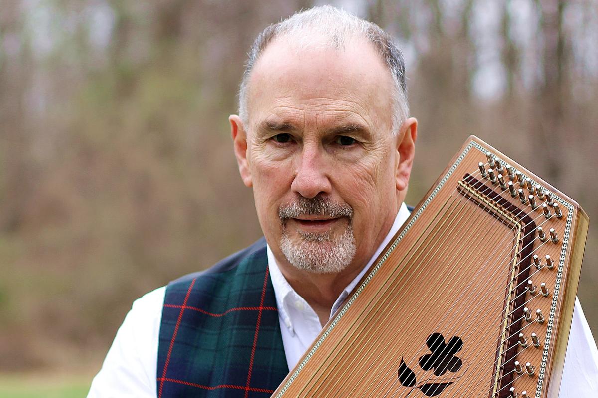 A man holding an stringed instrument in a white dress shirt and plaid vest against a blurred wooden background 