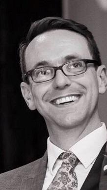 Black and white photo of a white man, dressed in suit and tie with glasses