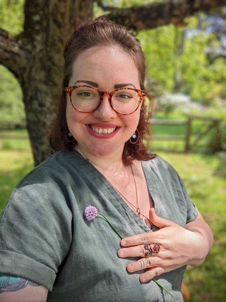 A woman in a green gray shirt  holding a flower against a background of a tree and greenery. 