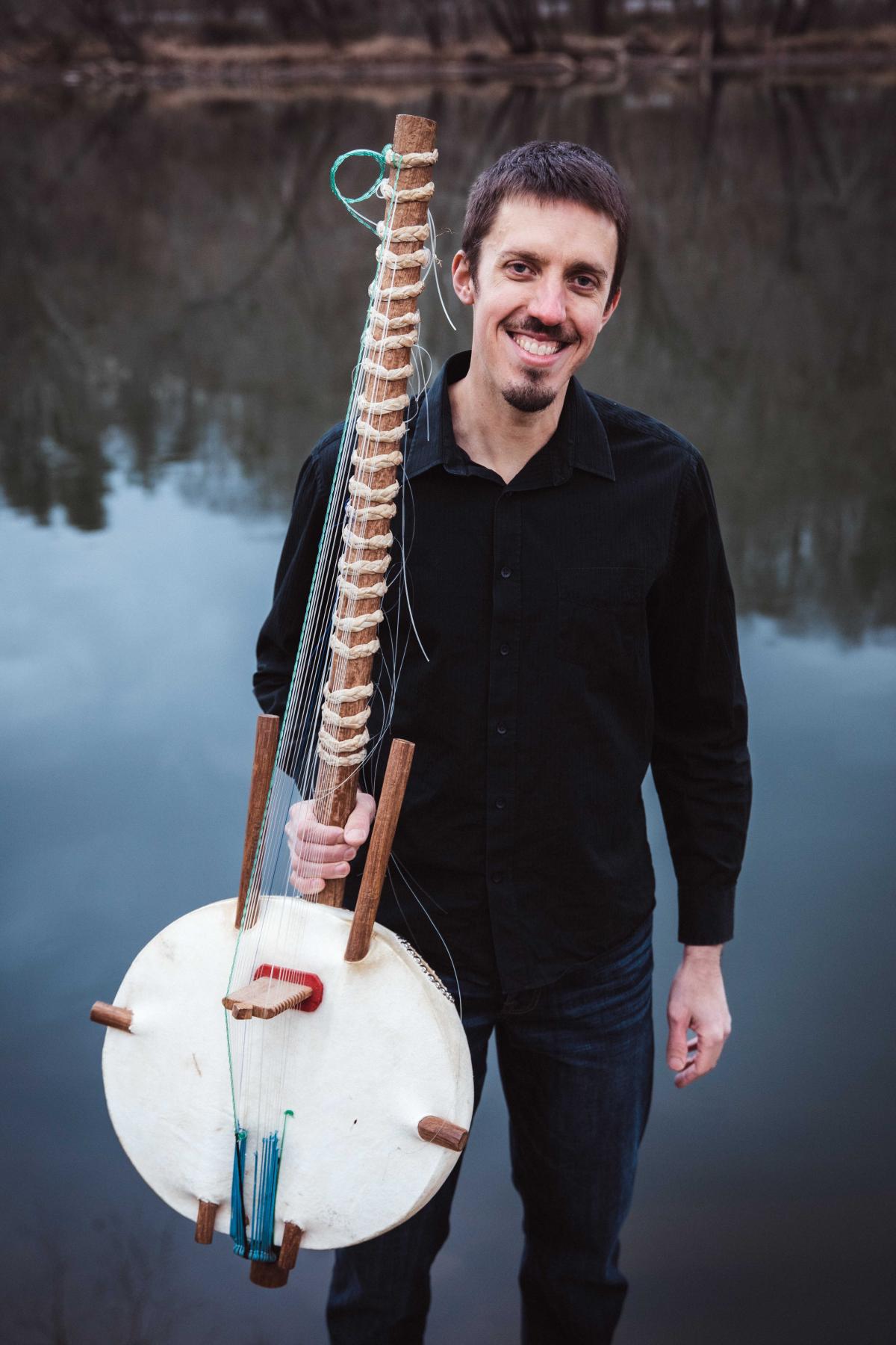 a man stands in front of a lake holding a Kora, with a black shirt and dark jeans.