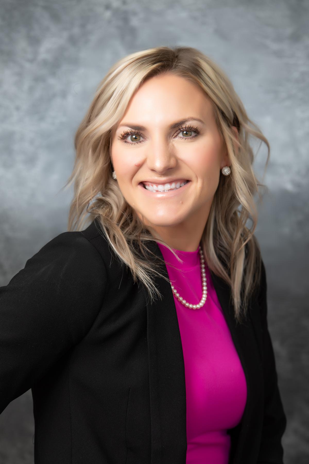 Kara Miller looking into the camera and smiling. She is wearing a hot pink shirt and black jacket. 