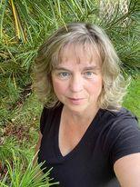 A person next to pine bushes looking into the camera with a black shirt